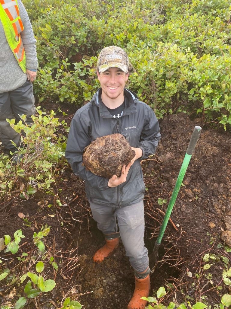 Liam Donovan, UBC Forestry Co-op student, standing in a pit holding a rock and smiling. 