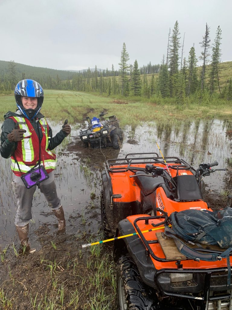 Hailey standing in the mud beside an ATV wearing driving gear.