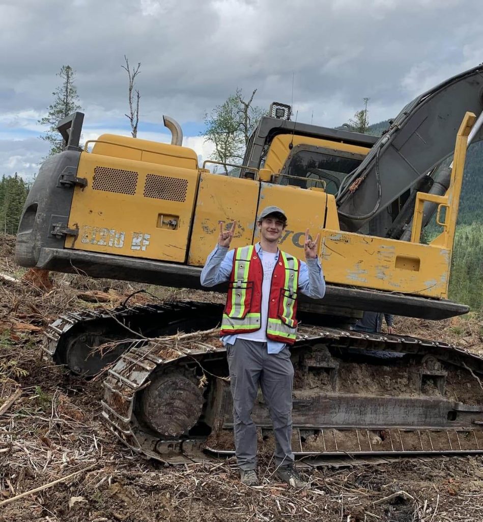UBC Forestry Co-op Student, Matthew Appelbum, standing in front of a tractor in the forest and smiling. 