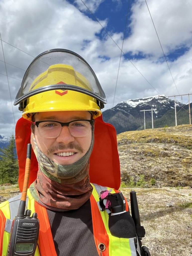 Nicholas Mantegna (a UBC Forestry Co-op student) standing near a mountain, wearing safety gear and smiling